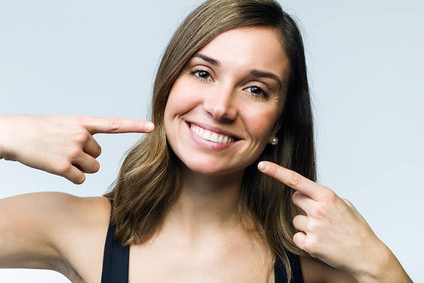 Beautiful young woman pointing to her perfectly white smile. If you’ve been duped by an over-the-counter treatment, or you are finally ready to transform your smile, the team at Irresistible Smiles can help!